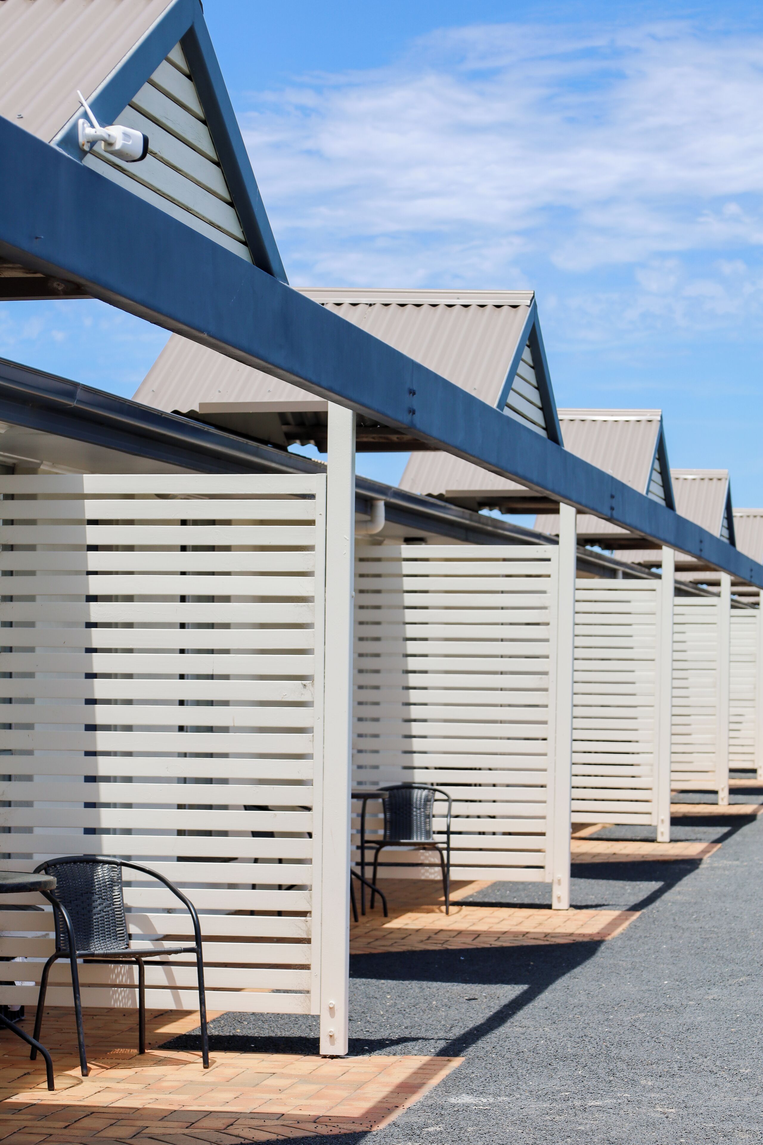 A row of white, slatted room walls with a metal roof structure, a paved walkway, and two metal chairs.