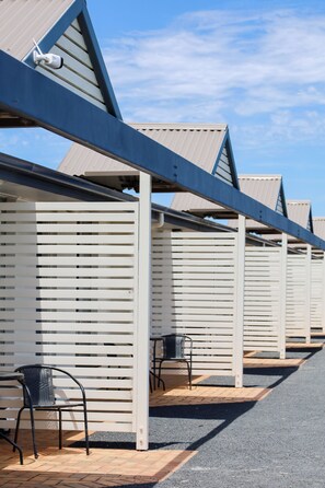 A row of white, slatted room walls with a metal roof structure, a paved walkway, and two metal chairs.