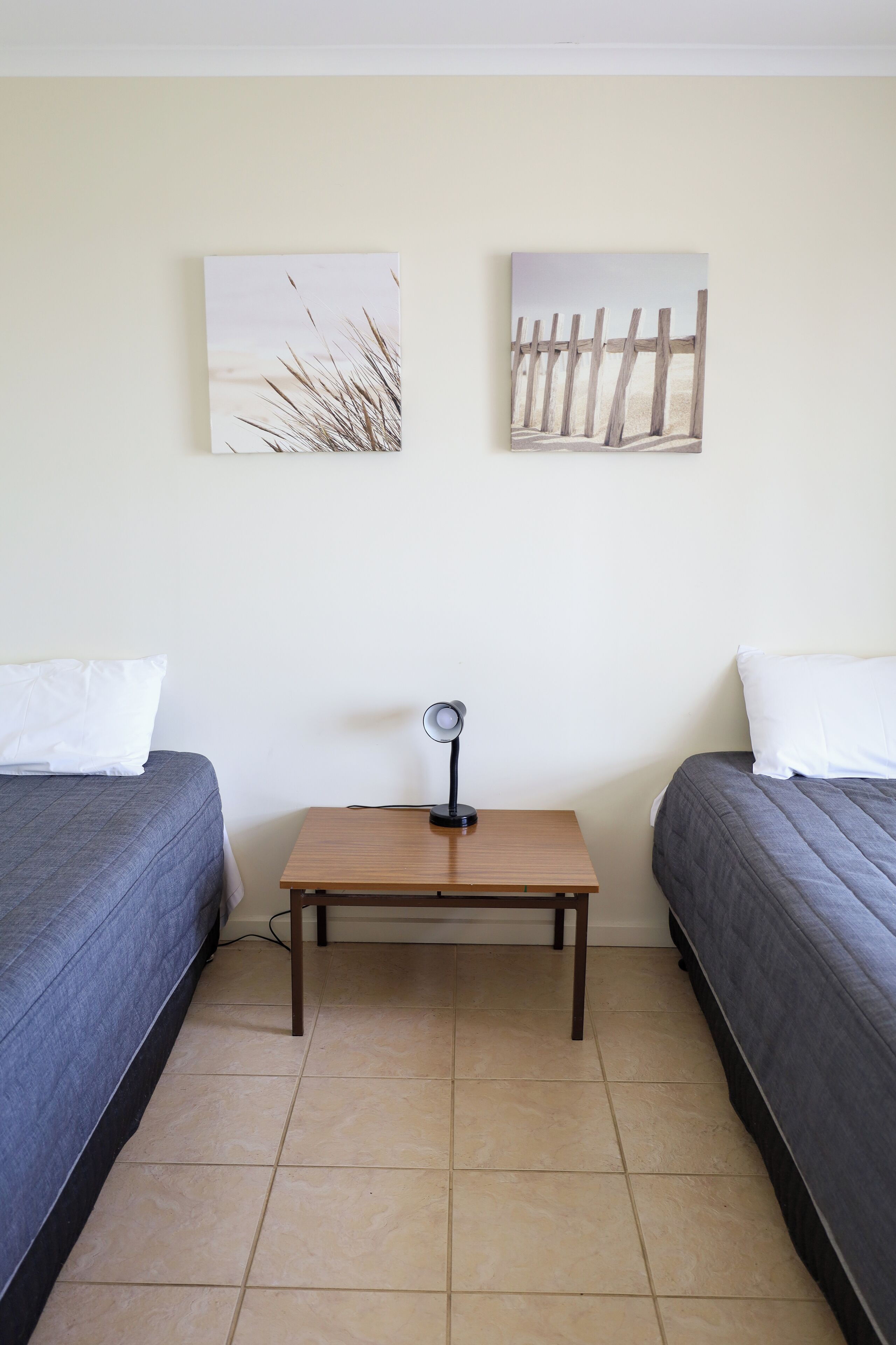 A room with two beds, a small table with a lamp, and two wall-mounted paintings.