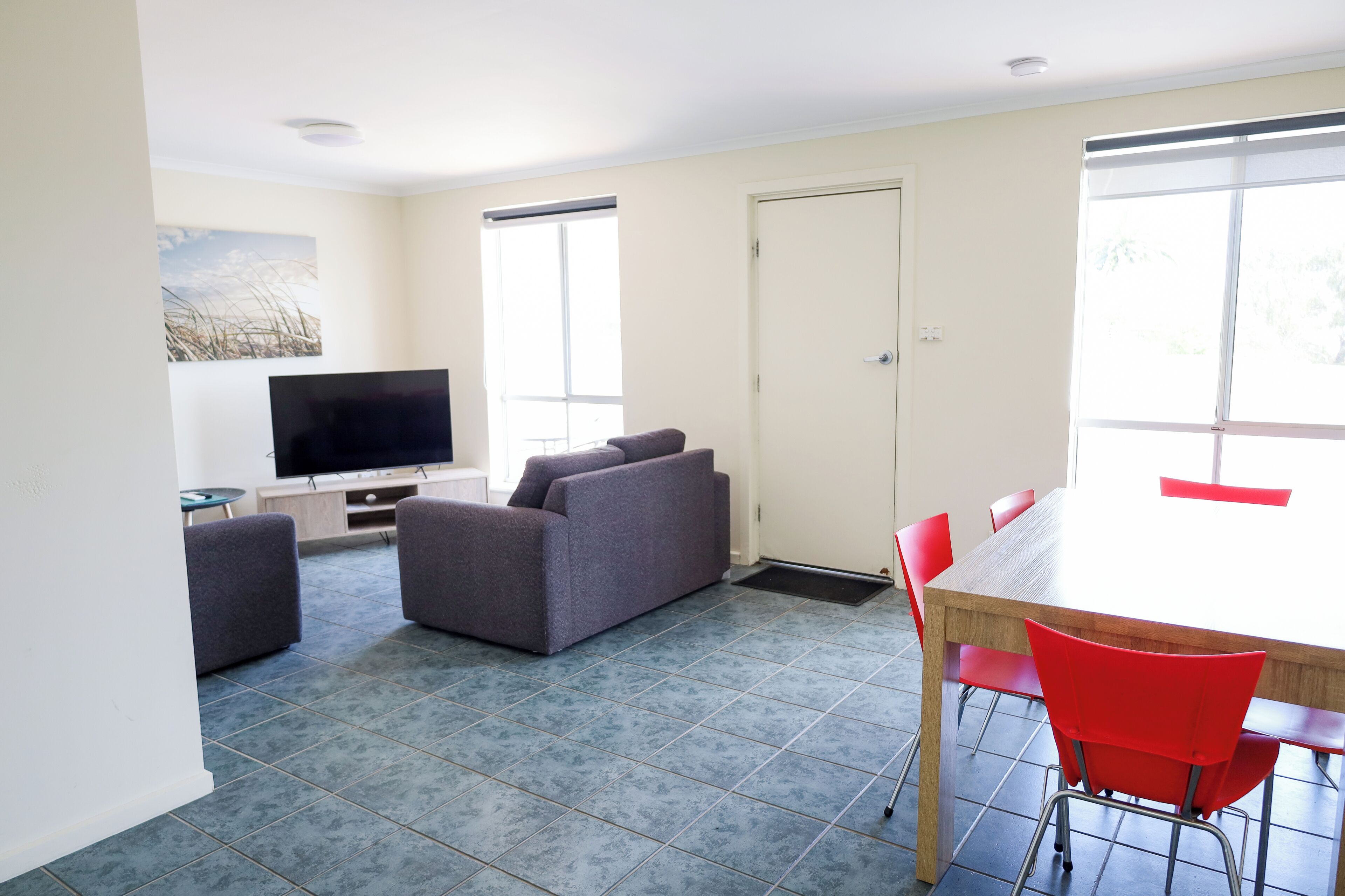 A room with a flat-screen TV, grey sofas, a dining table with red chairs, and a large window.