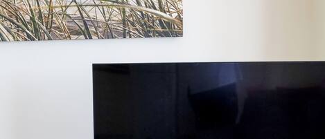 Un televisor de pantalla plana montado en un mueble de madera, una mesa de vidrio con un mantel individual azul y una silla en una habitación con suelo de baldosas.
