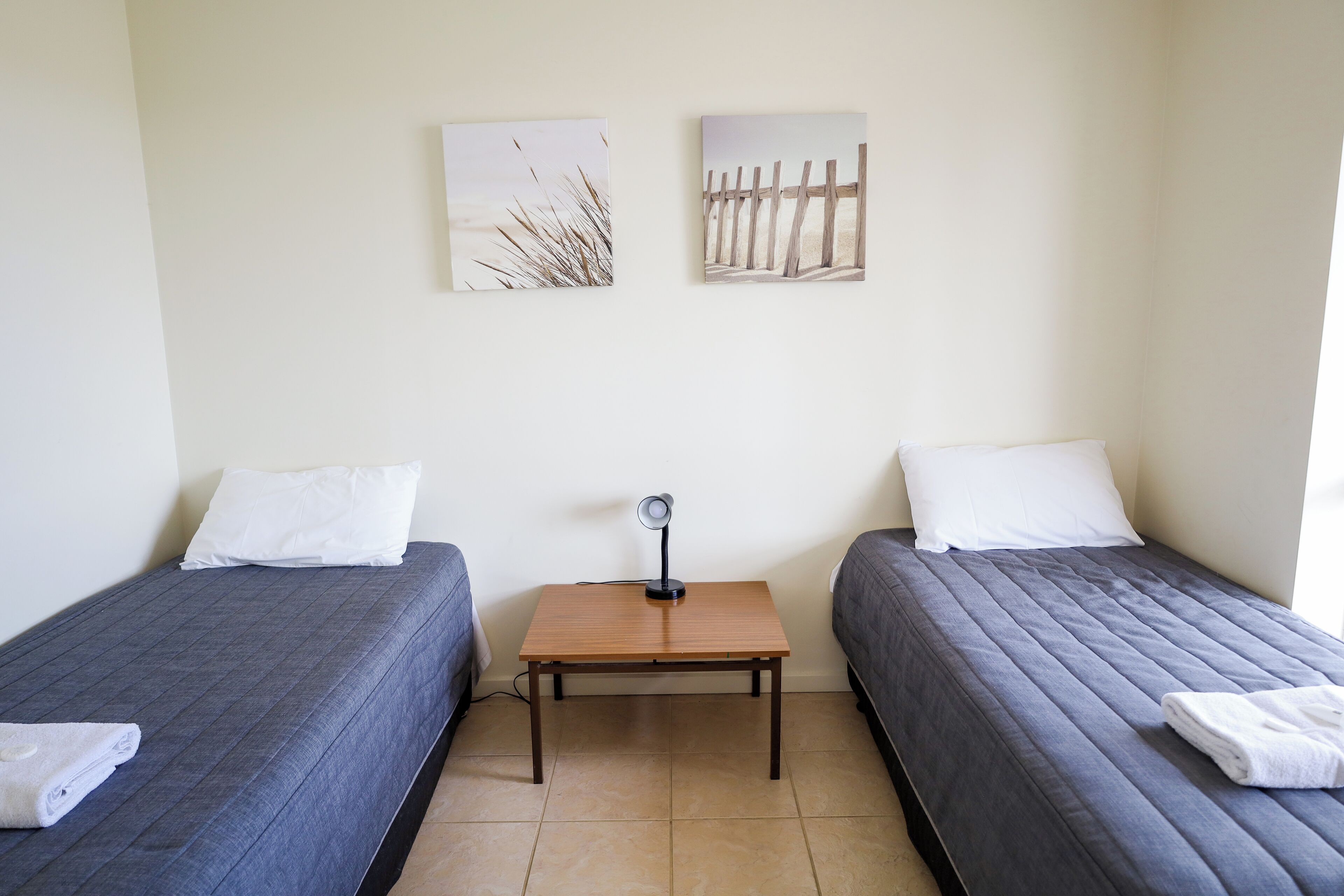Une chambre avec deux lits, une petite table avec une lampe et deux tableaux accrochés au mur.