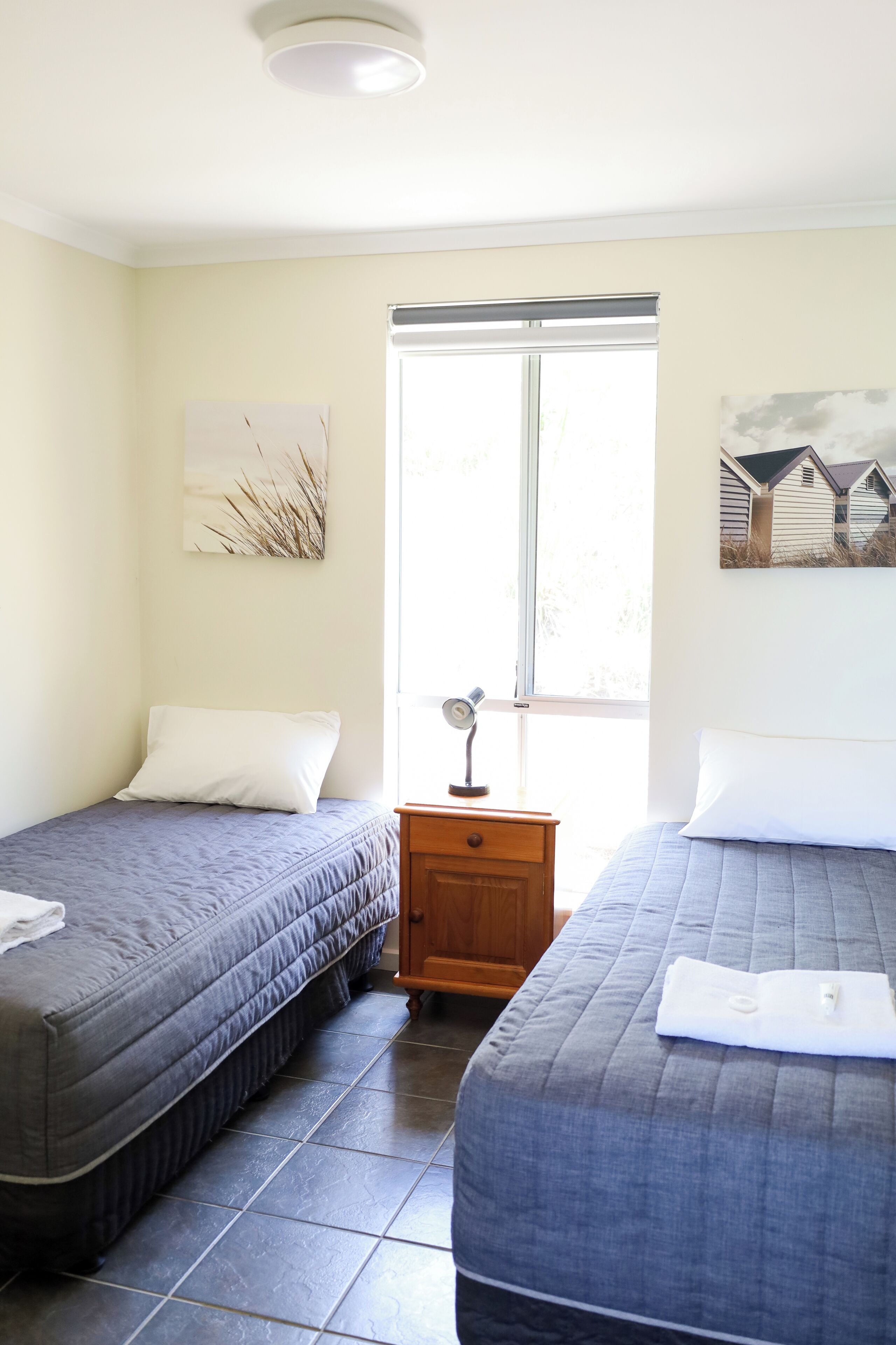 Ein Zimmer mit zwei Betten, einem Fenster, einem Nachttisch und Wandbildern.