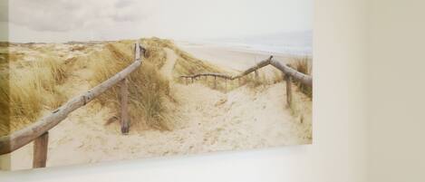 A bed with white pillows and a gray quilt, a wall-mounted picture of a beach scene, and a black lamp on the right.