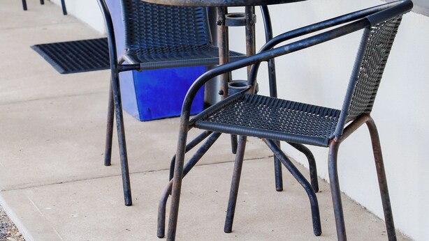 A glass-top table with two wicker chairs, a potted plant, and a blue trash bin.