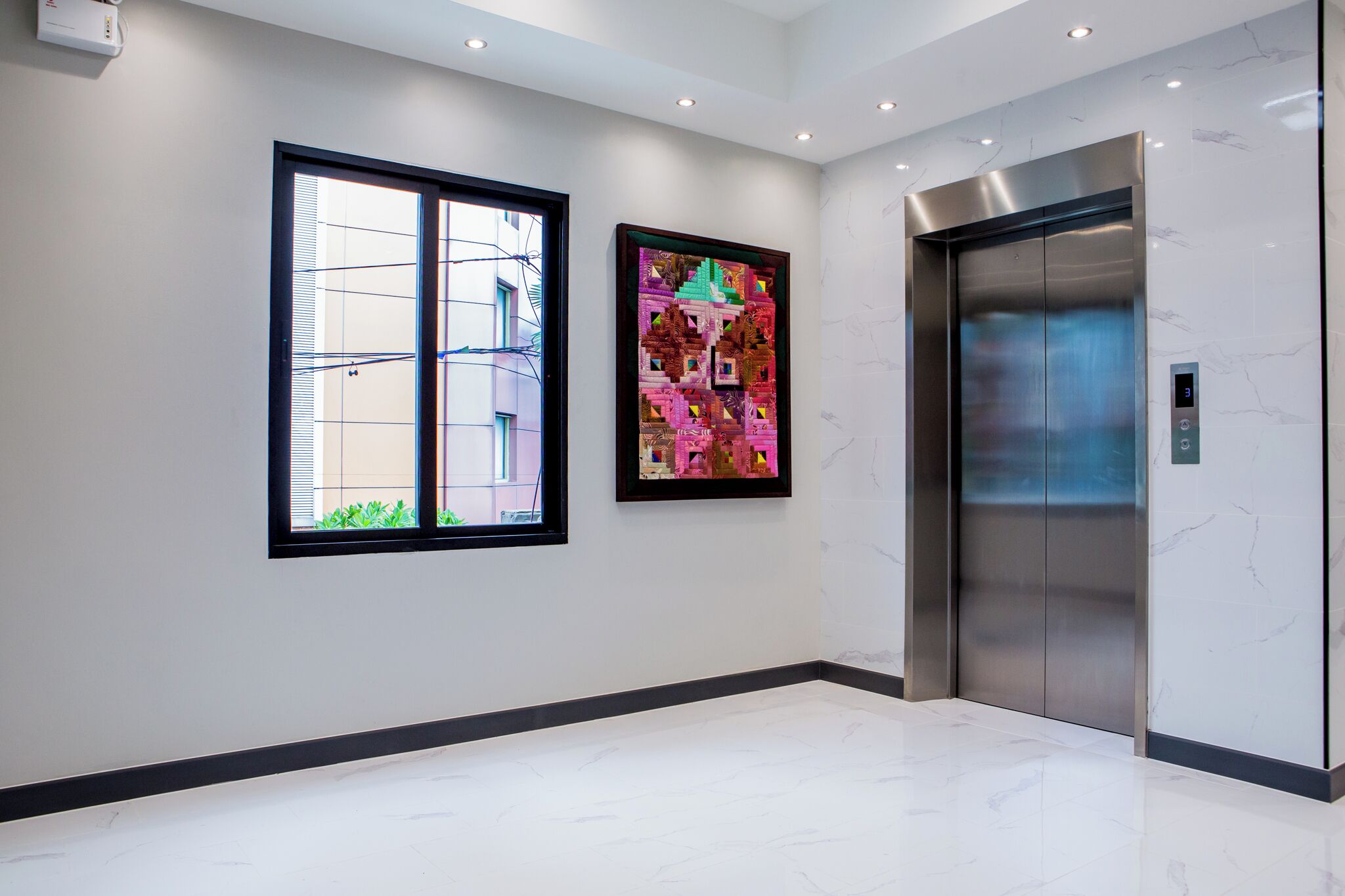 A modern elevator with a digital display, a framed colorful artwork on the wall, and a window with blinds.