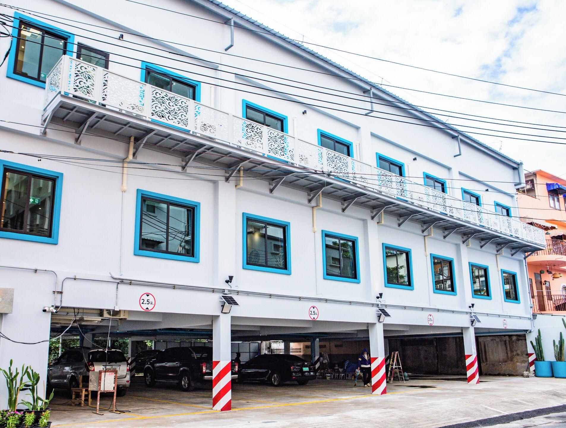 A multi-story building with a white facade and blue window frames, featuring a covered parking area with several cars and a person walking.