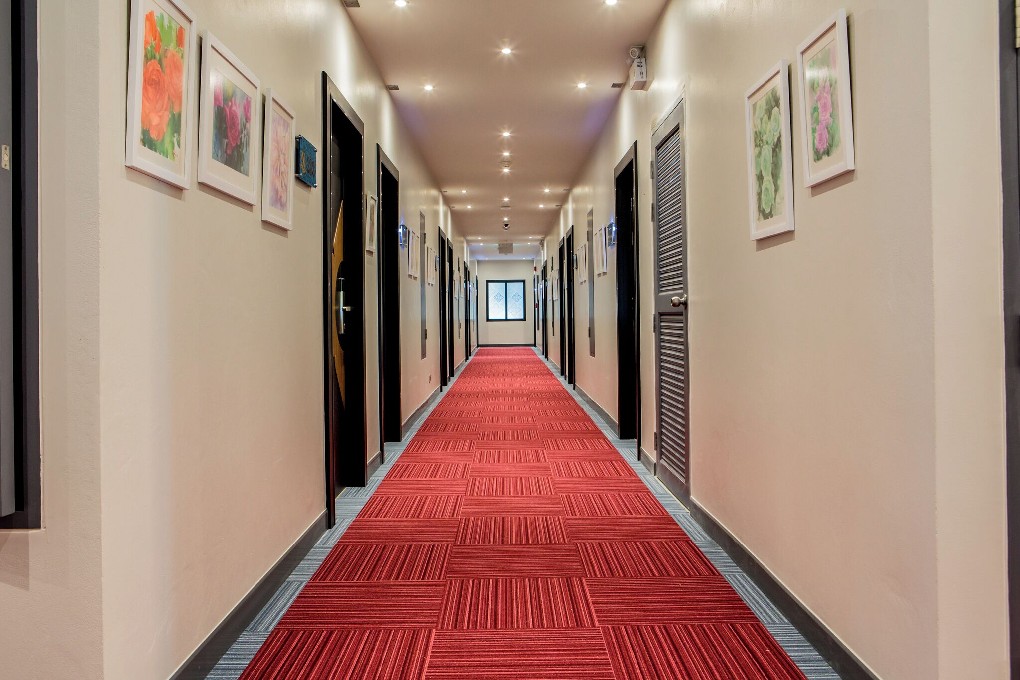 A hotel hallway with a red carpet, framed floral artwork on the walls, and doors leading to rooms.