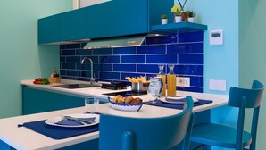 A modern kitchen with teal cabinetry, a white countertop, and a breakfast setup including a bowl of fruit, a bottle of orange juice, and a coffee maker.