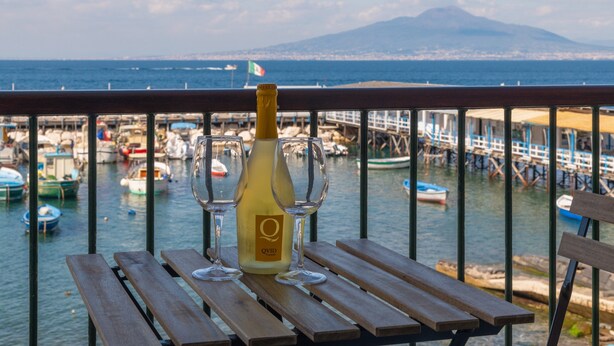 A bottle of QVID wine and two glasses on a wooden table with a view of boats and a distant mountain.