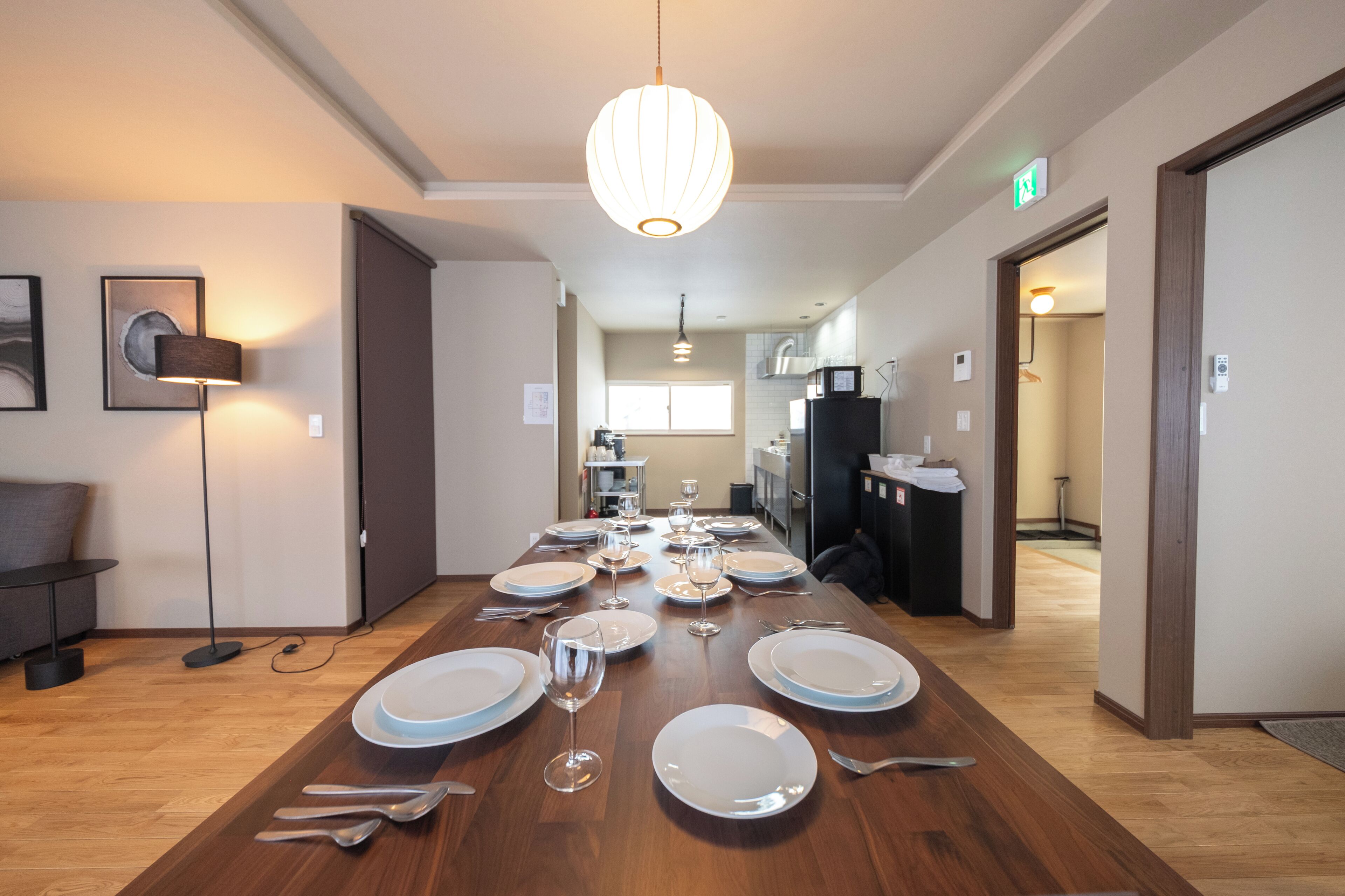 A dining area with a wooden table set for a meal, a hanging light fixture, and a kitchen in the background.