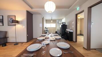 A dining area with a wooden table set for a meal, a hanging light fixture, and a kitchen in the background.