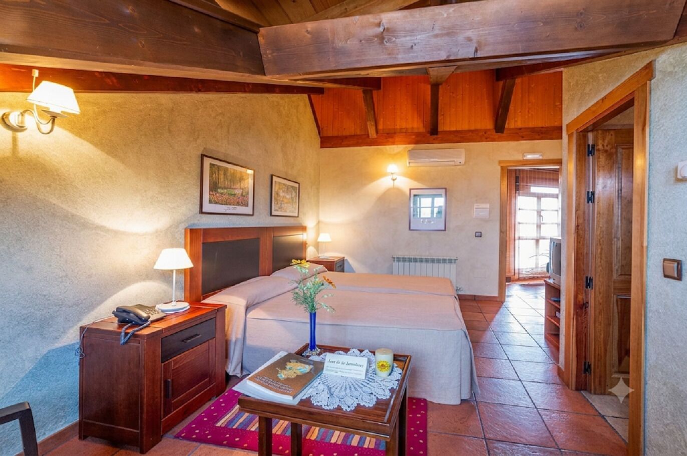 Una habitación de hotel acogedora, con techo de madera, una cama, un escritorio con teléfono, una mesita con un libro y un florero, y un radiador.