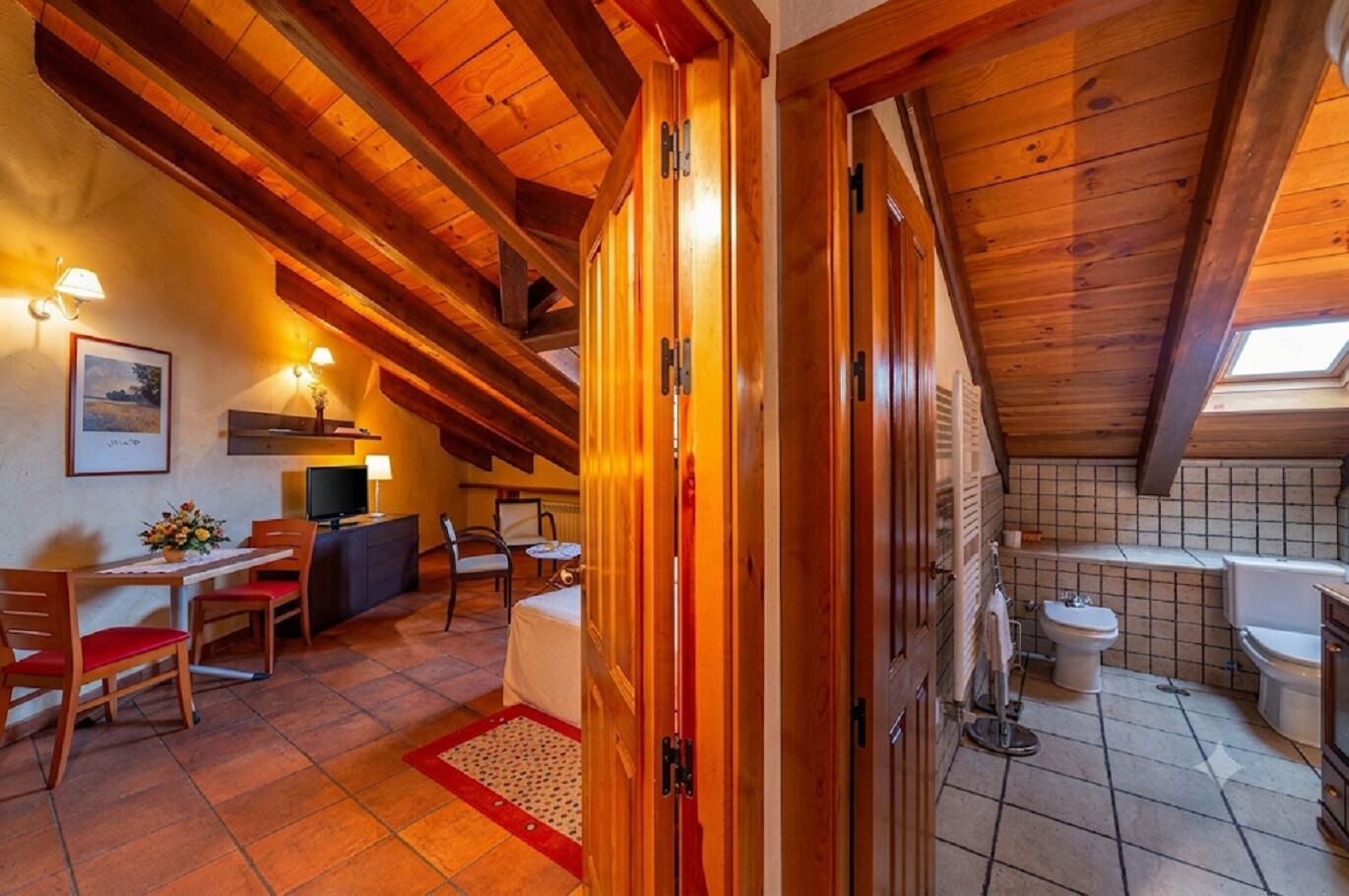 Una habitación acogedora con techo de madera, un comedor con sillas y una mesa, un televisor pequeño y un baño con inodoro y lavamanos.