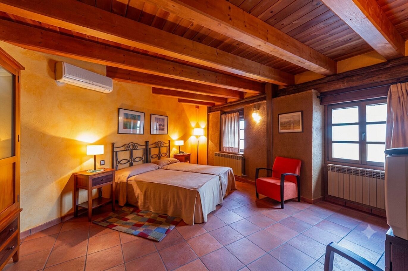 Un dormitorio con cama, mesitas de noche, una silla roja, una ventana y un radiador.