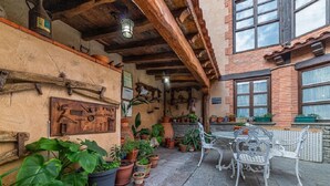 A rustic outdoor patio with stone flooring, potted plants, a metal table with chairs, and a wooden wall with various tools and decorations.