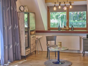 Una habitación acogedora con un aparador de madera, una mesa con libros, una silla y una ventana con vista a los árboles.