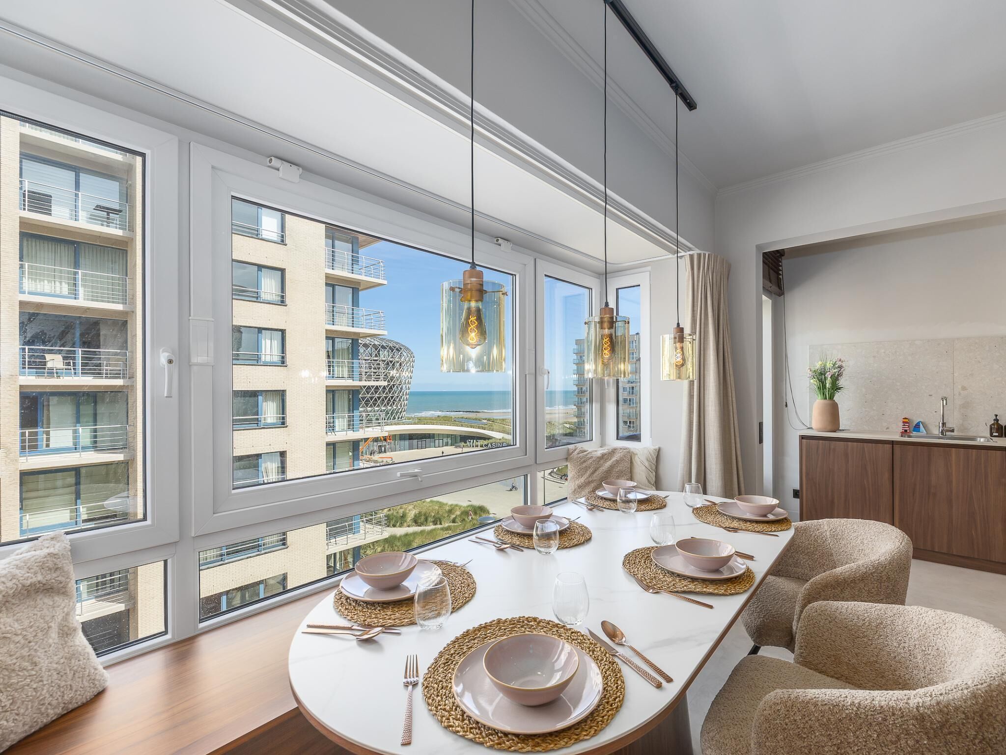 Een moderne eetkamer met een grote tafel gedekt voor vier personen, grote ramen en uitzicht op een gebouw en strand.
