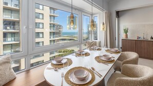 Ein moderner Essbereich mit einem großen Tisch für vier Personen, großen Fenstern und Blick auf ein Gebäude und einen Strand.