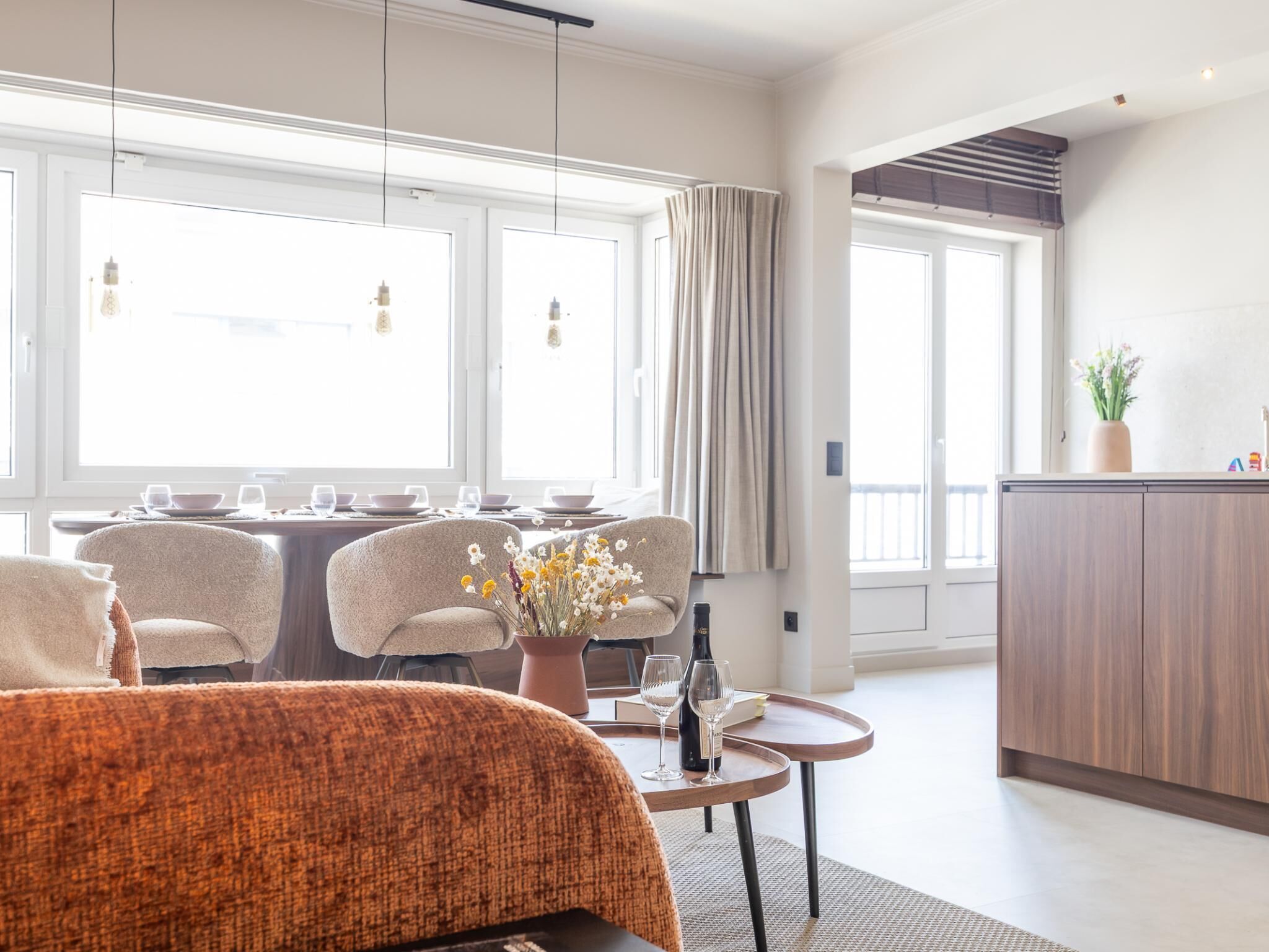 A modern hotel room with a bed, a dining area with chairs and a table, a small kitchen area, and large windows.