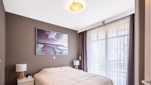 Une chambre d’hôtel avec un lit, une table de chevet, une lampe, un tableau et une fenêtre avec des rideaux.