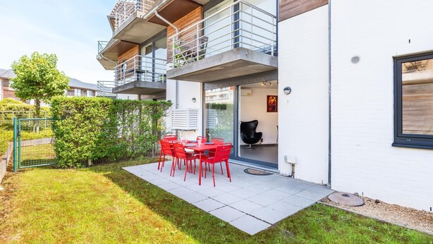 Un edificio moderno de dos pisos con un balcón, un patio con sillas rojas y vistas del entorno.