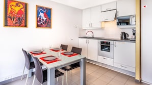 Una cocina moderna con armarios blancos, un microondas, un horno y un fregadero. Hay una mesa de comedor con sillas y salvamanteles rojos. Dos obras de arte enmarcadas están en la pared.