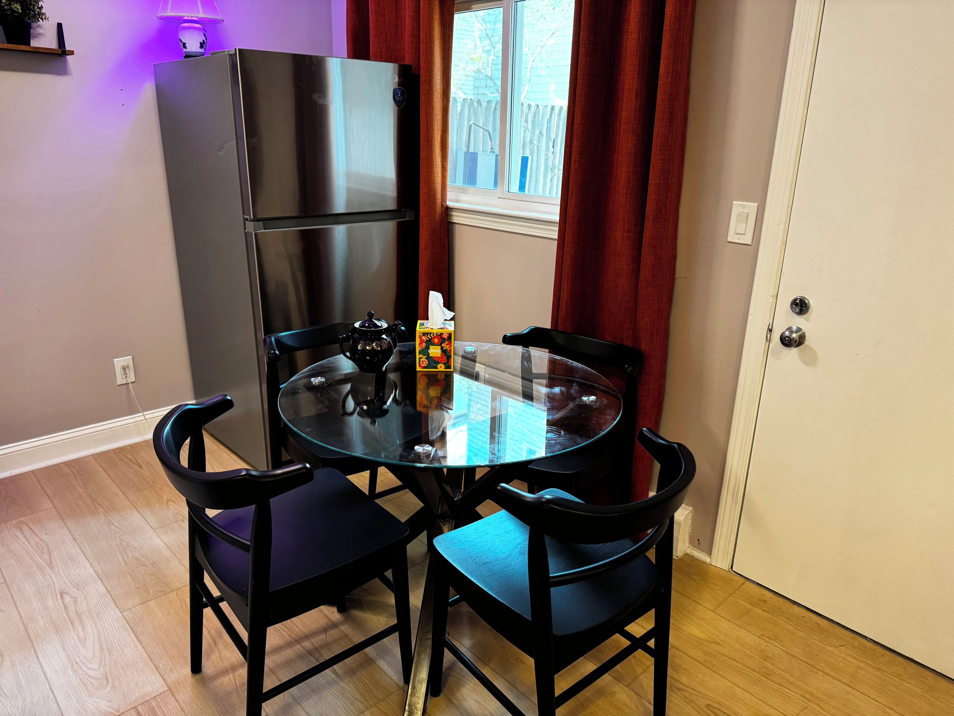 A compact dining area with a glass table, four black chairs, a black refrigerator, and a window with red curtains.