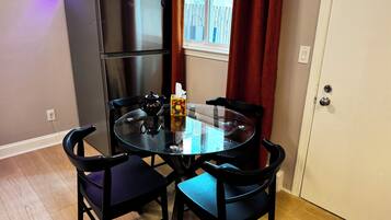 A compact dining area with a glass table, four black chairs, a black refrigerator, and a window with red curtains.