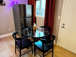 A compact dining area with a glass table, four black chairs, a black refrigerator, and a window with red curtains.