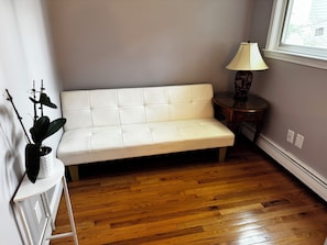 A room with a white sofa, a wooden floor, a lamp, and a window.