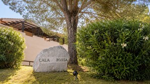 Um letreiro de pedra com a inscrição 'CALA ROSSA', uma árvore e uma sebe bem cuidada.