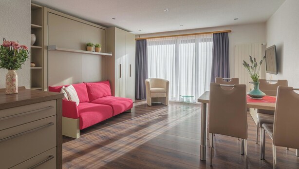 Moderni hotellihuone, jossa on sohva, ruokapöytä, tuoleja, televisio ja ikkuna, jossa on verhot.