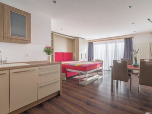 A hotel room with a bed, a desk, a chair, a small table, a vase with flowers, and a window with curtains. - Alpen Leukerbad # 2-bed apartment, Dusche, WC by Interhome (Leukerbad)