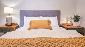Una cama bien hecha con cabecera, dos mesitas de noche con lámparas, un plaid tejido y una planta en maceta sobre la mesita.