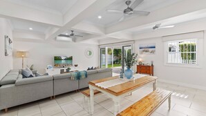 A spacious living room with a grey sofa, a wooden dining table, and ceiling fans. - Coastal Corner Poolside Retreat on The Strand (North Ward)