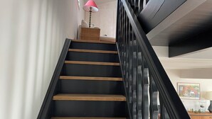 Una escalera con escalones negros y barandillas de madera que lleva a un rellano con una cesta de mimbre y una lámpara rosa.