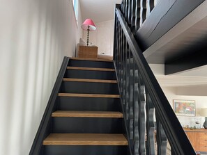 Eine Treppe mit schwarzen Stufen und Holzhandläufen, die zu einer Zwischenebene mit einem Korbwägelchen und einer rosa Lampe führt.