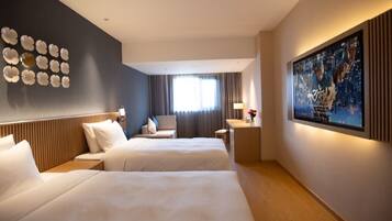 A hotel room with two beds, a desk, a chair, a window with curtains, a wall-mounted artwork, and a ceiling light.