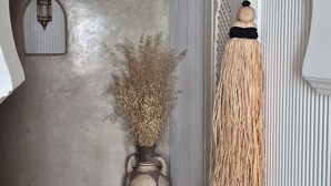 Un arco d'ingresso conduce a una stanza con parete strutturata, una lanterna sospesa, un vaso con piante essiccate e una scopa appoggiata al muro.