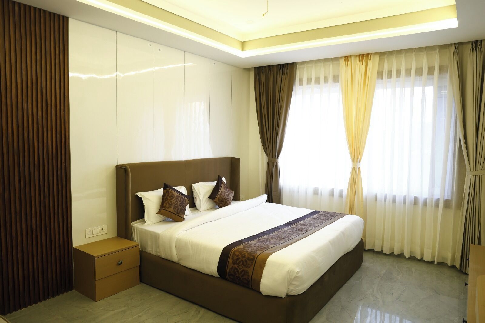 Quarto de hotel moderno com uma cama grande, uma mesa de cabeceira, parede de painéis de madeira e uma janela com cortinas.