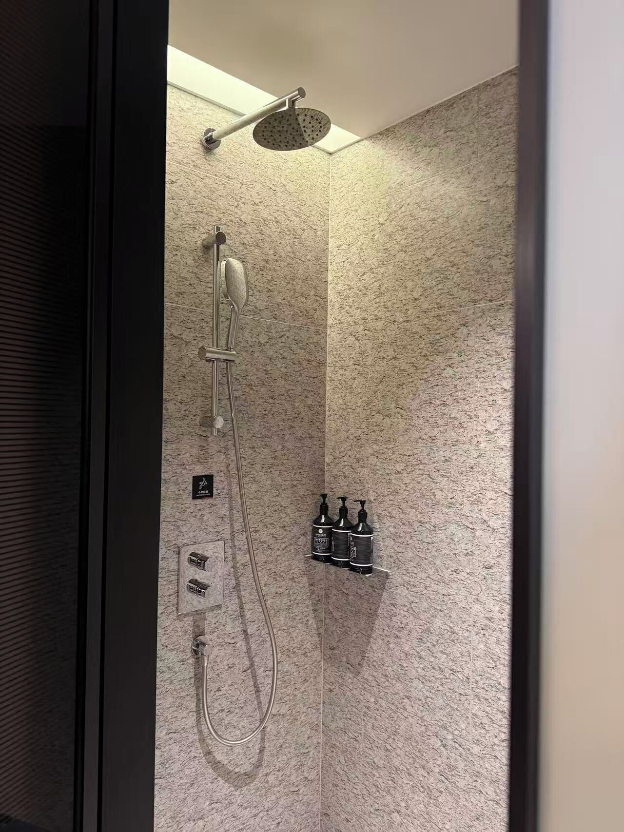 Une douche équipée d’un pommeau de douche à main, d’un pommeau de douche fixé au mur et d’un pommeau de douche à pluie à main. Trois bouteilles de shampoing et d’après-shampoing sont posées sur une étagère.