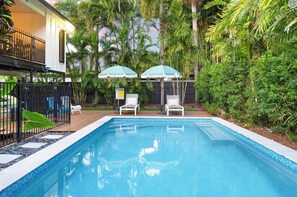 A swimming pool with two lounge chairs and umbrellas, surrounded by lush greenery and a wooden deck. - Tropical Poolside Studio (Mundingburra)