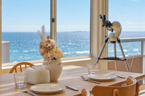 A dining table with a vase of flowers, plates, cutlery, and a telescope on a balcony with a city and ocean view. - Pudding's Home: Exclusive Oceanfront Experience (Frankston)