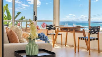A modern living room with a large window overlooking the ocean, a dining area with a wooden table and chairs, and a sofa with decorative pillows.