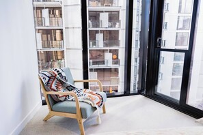 Ein Eckzimmer mit Holzstuhl, einem gemusterten Überwurf und einem großen Fenster mit Blick auf ein Gebäude.