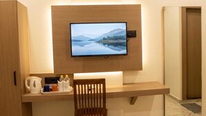 Habitación de hotel con televisor de pantalla plana montado en una pared de madera, una silla de madera, una mesita con tetera, tazas y botellas.