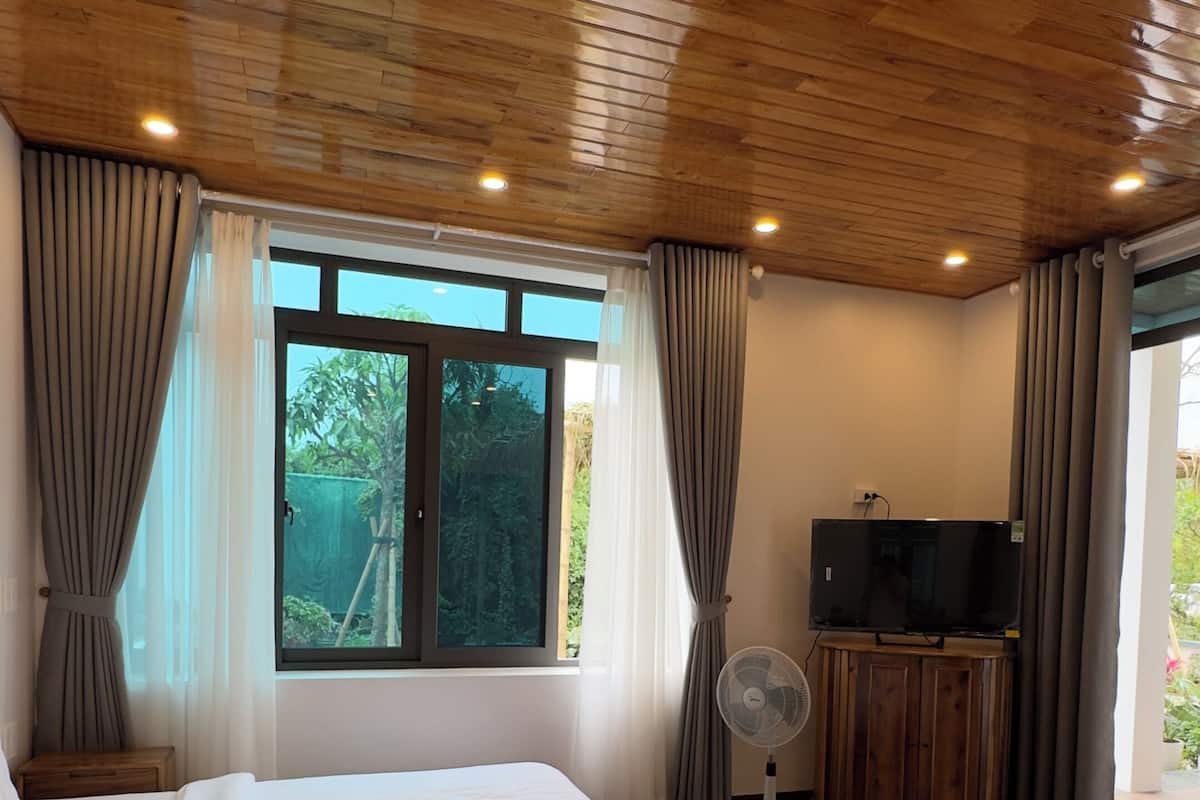 Una habitación con piso de madera, dos camas, un televisor, un ventilador y una ventana con cortinas.