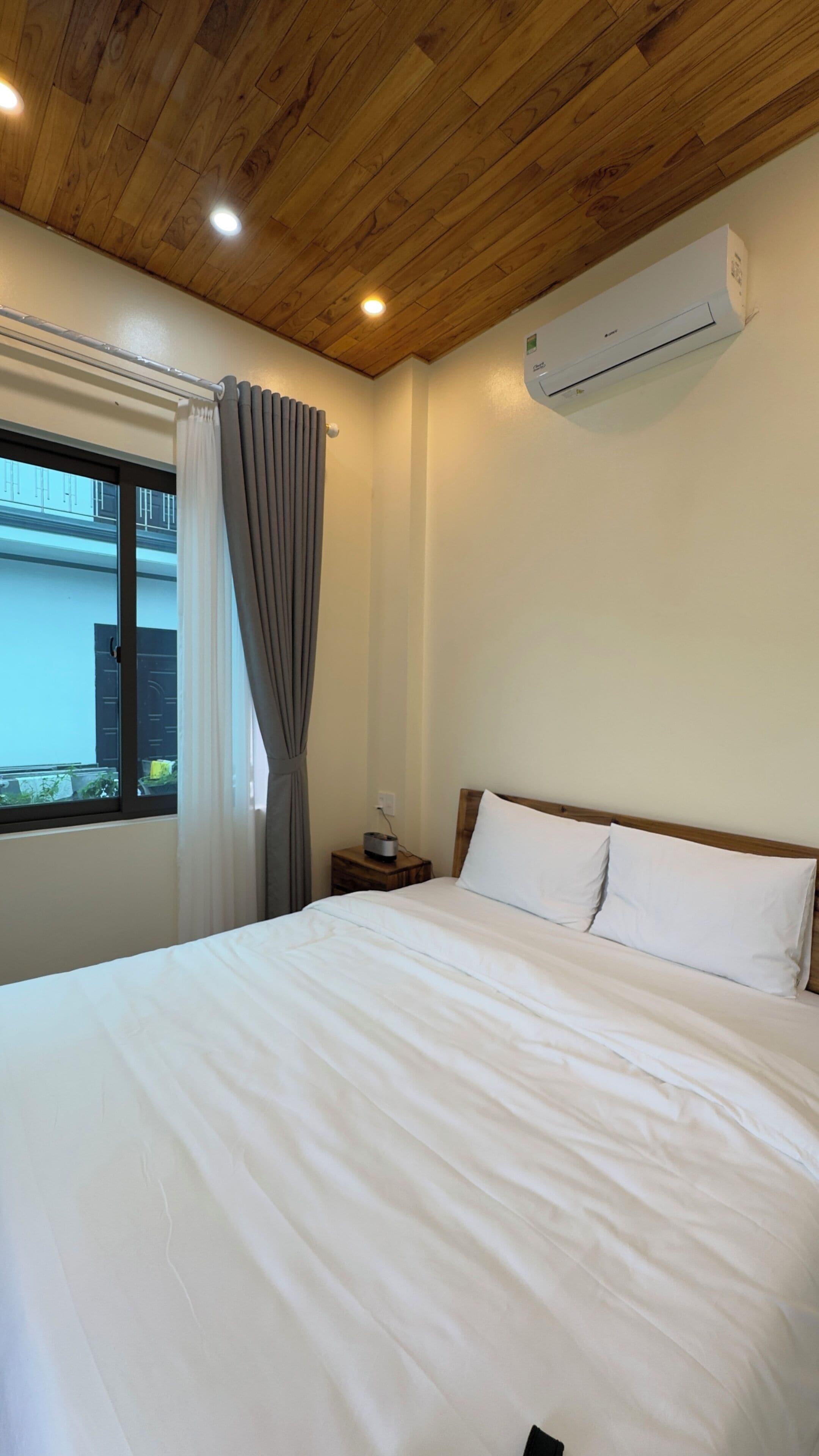 Una camera da letto con soffitto in legno, un letto con biancheria bianca, una finestra con tende e un condizionatore d'aria.