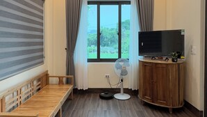 A room with a wooden ceiling, a large window with curtains, a wooden bench, a TV on a wooden cabinet, and a standing fan.